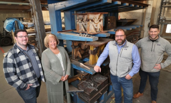 Four people stand in a metalworking shop beside blue racks filled with large wooden and metal molds.