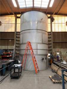 FAB Large metal cylindrical tank inside an industrial workshop, with an orange ladder leaning against it and tools and equipment around.
