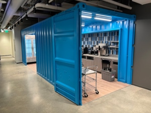 INSTALL Bright blue shipping container repurposed as a kitchenette with coffee machines and a rolling cart inside an office hallway.